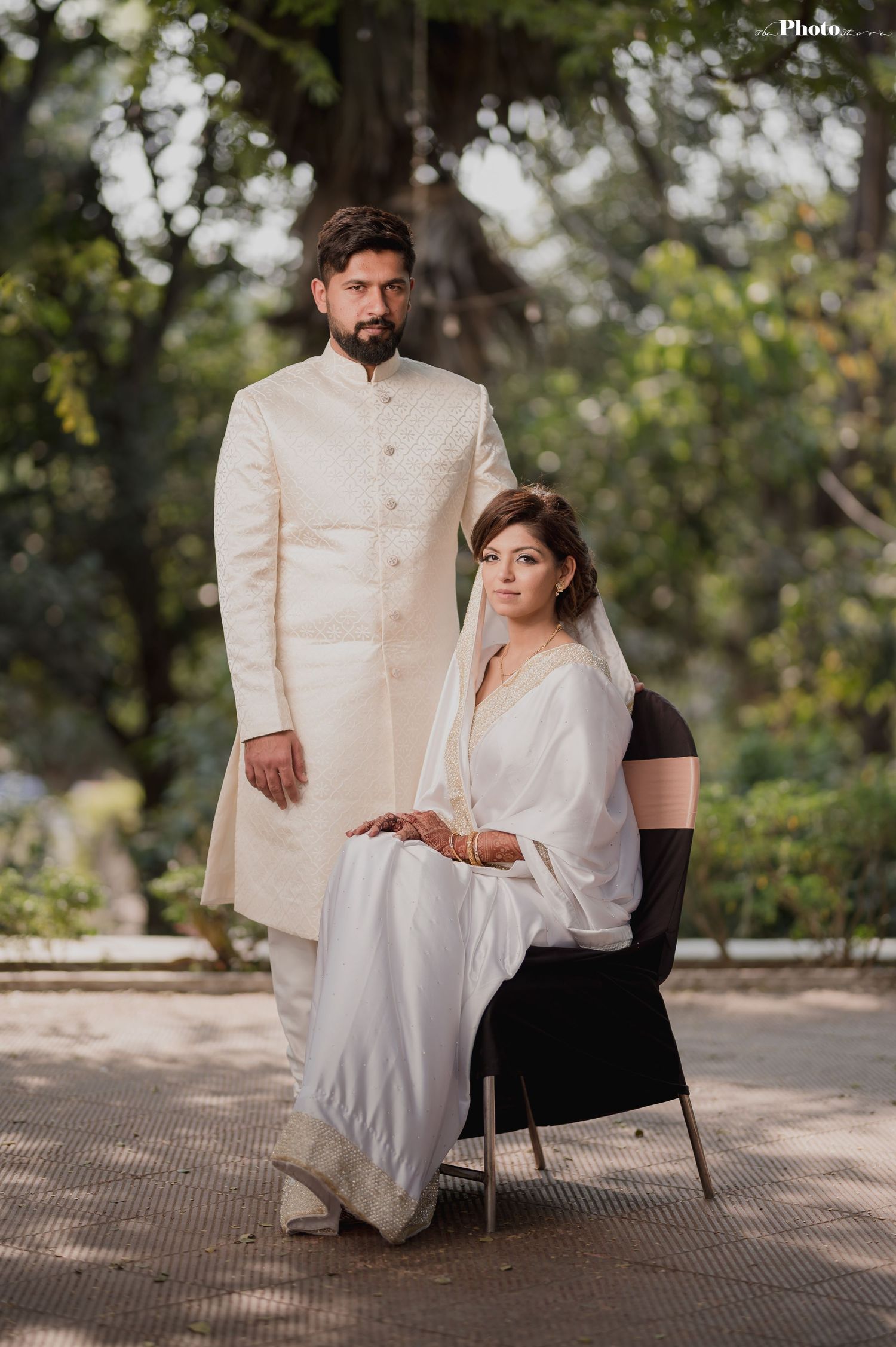 An elegant wedding portrait with the bride seated and groom standing in cream and white formal attire.