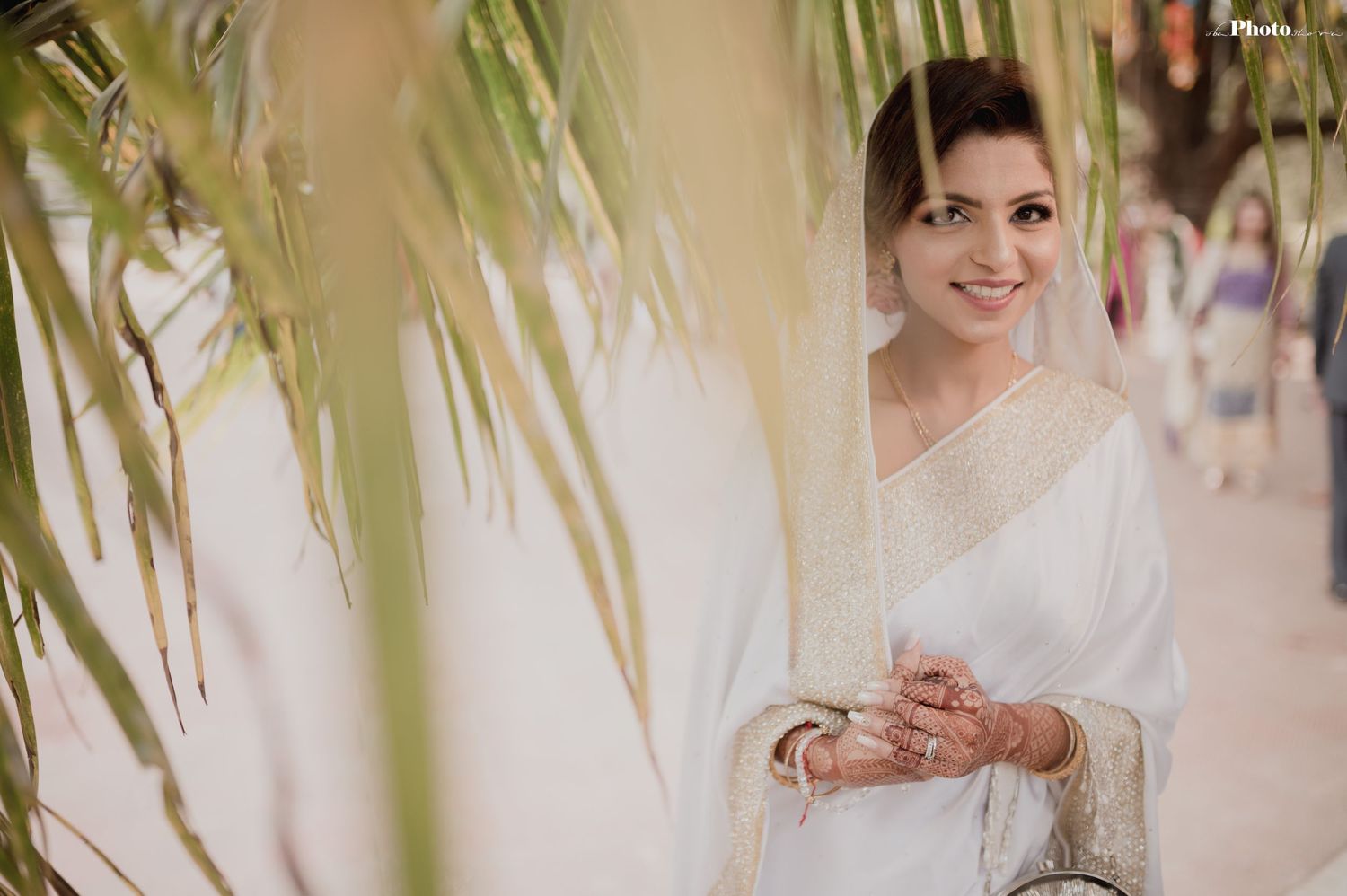 Bride in white ensemble shows traditional henna designs against palm leaf backdrop.