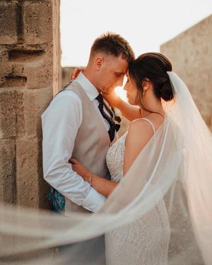 Intimate wedding moment captured in warm sunlight against stone wall with flowing veil.
