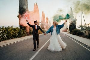 Wedding couple with colorful smoke bombs on palm-lined street creating festive atmosphere.