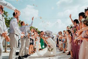 Wedding guests toss flower petals as newlyweds walk down aisle on sunny beach.