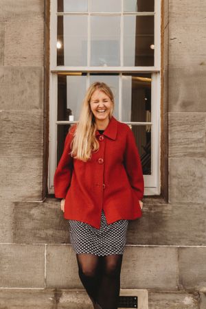 A joyful portrait against stone architecture wearing a bright red coat and polka dot skirt.