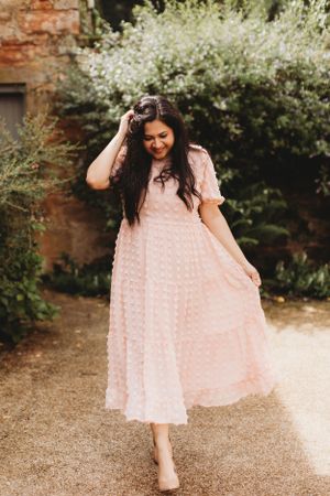 Woman in flowing pink dress twirls on garden path surrounded by blooming flowers.