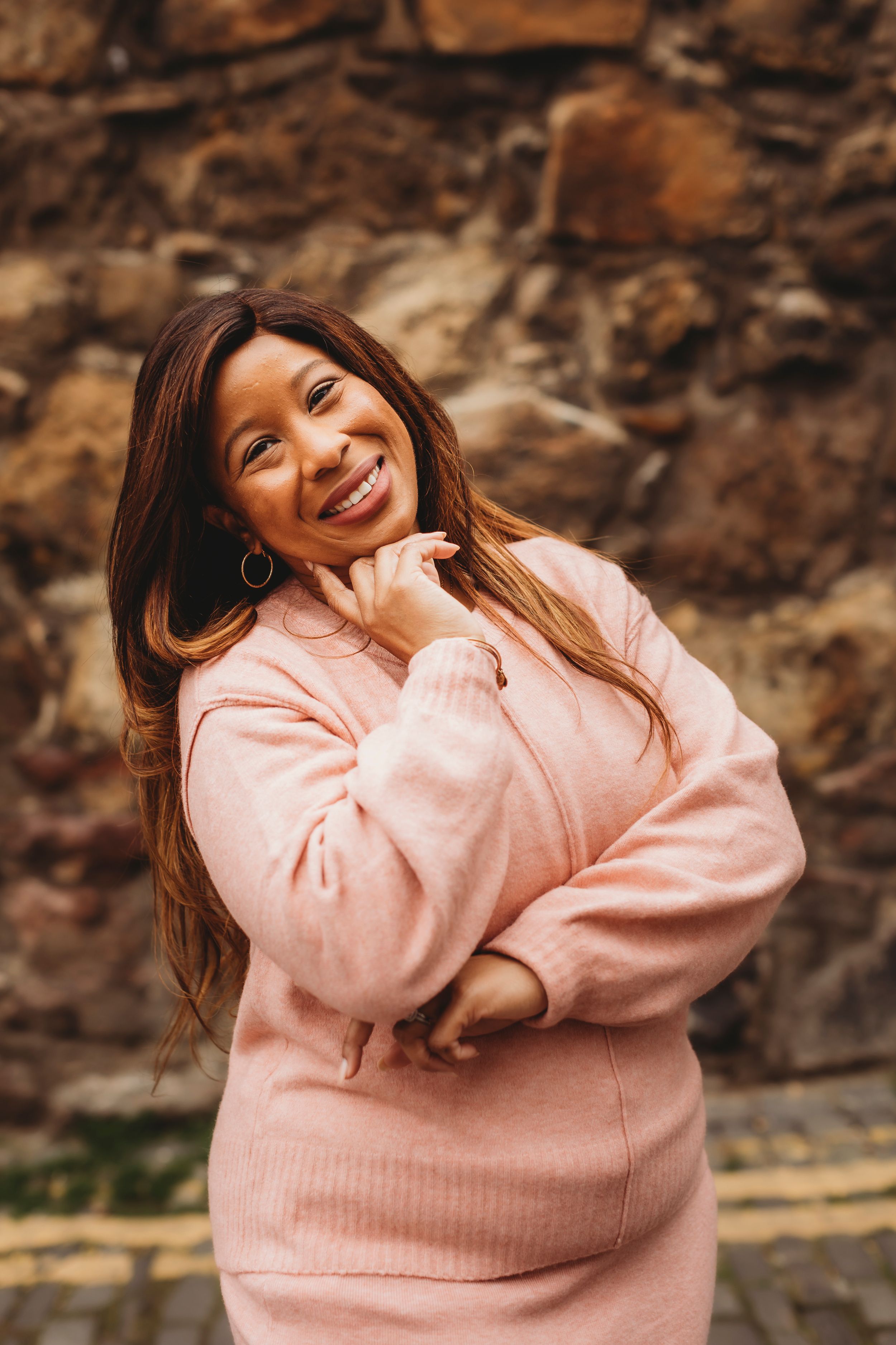 Woman in pink sweater smiling warmly against textured stone wall background.