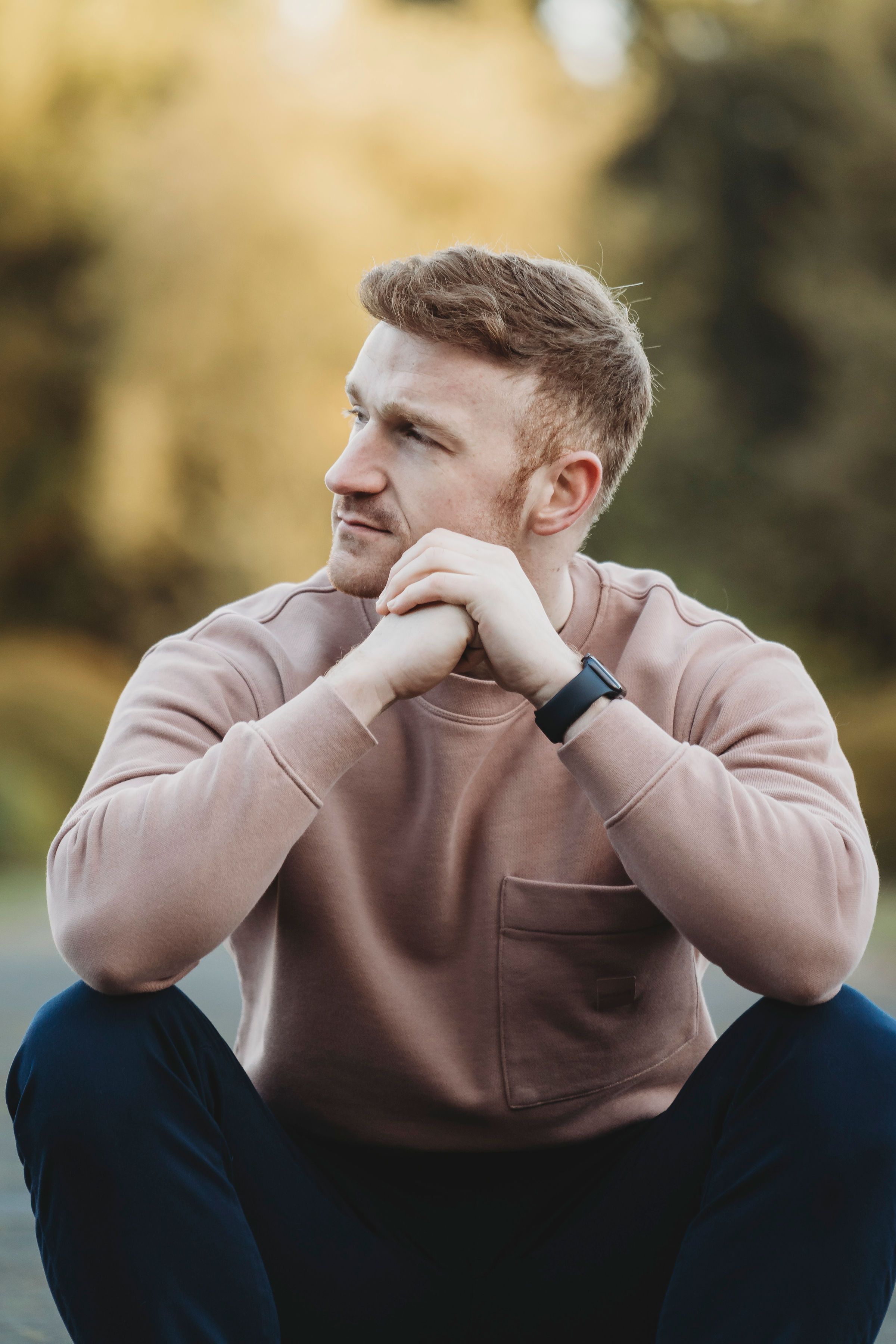 Thoughtful young person in beige sweatshirt sitting outdoors wearing a fitness tracker.