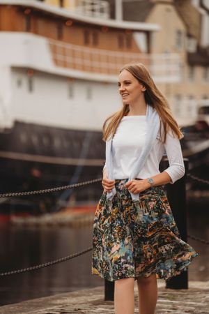 Person in white top and patterned skirt stands near docked ship on cloudy day.