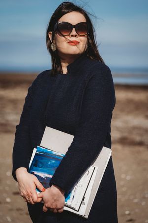 Woman in black coat holding books smiling at beach with sunglasses.