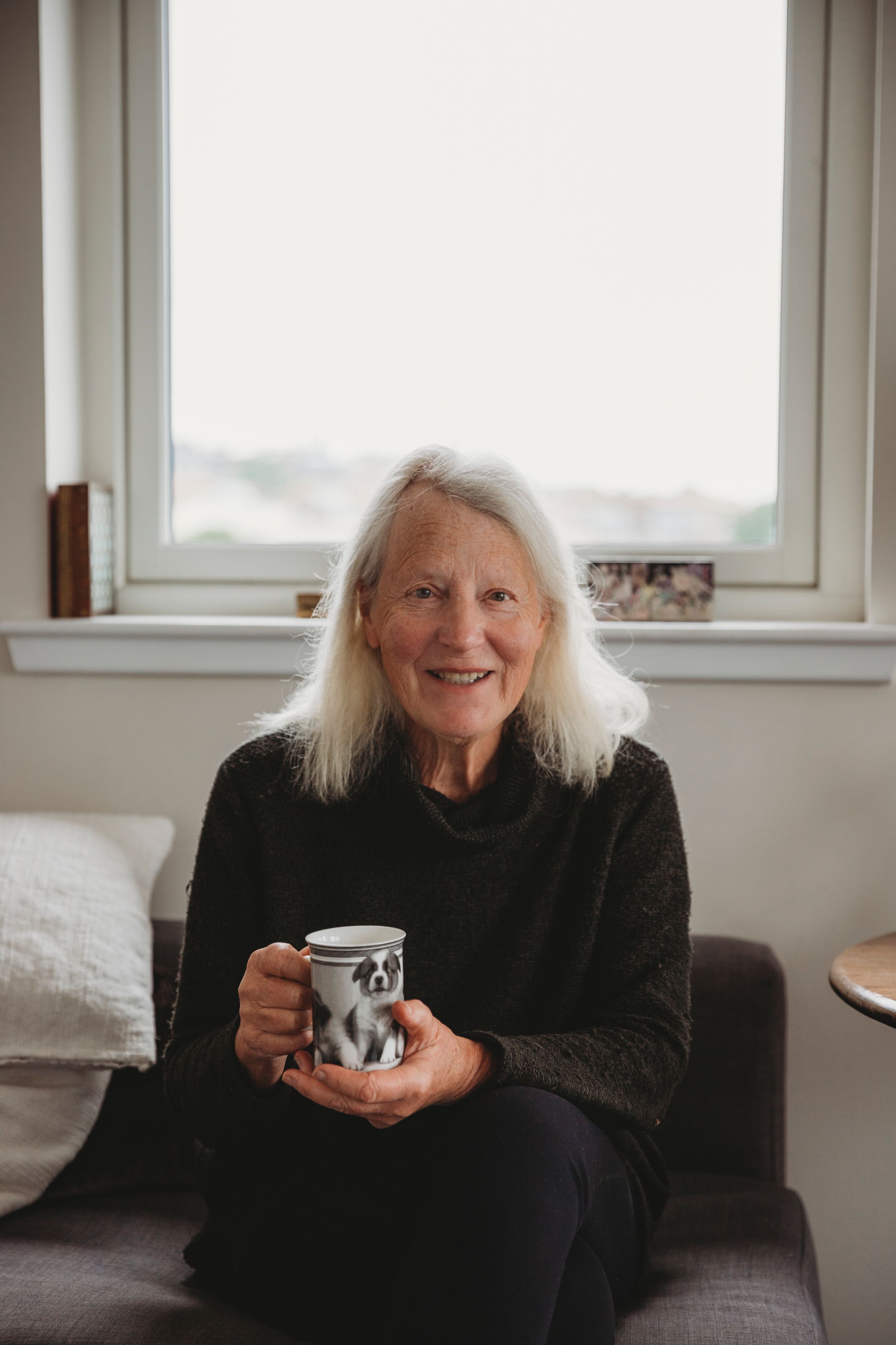 Someone relaxing on a couch by a window, holding a coffee mug and wearing dark casual clothing.