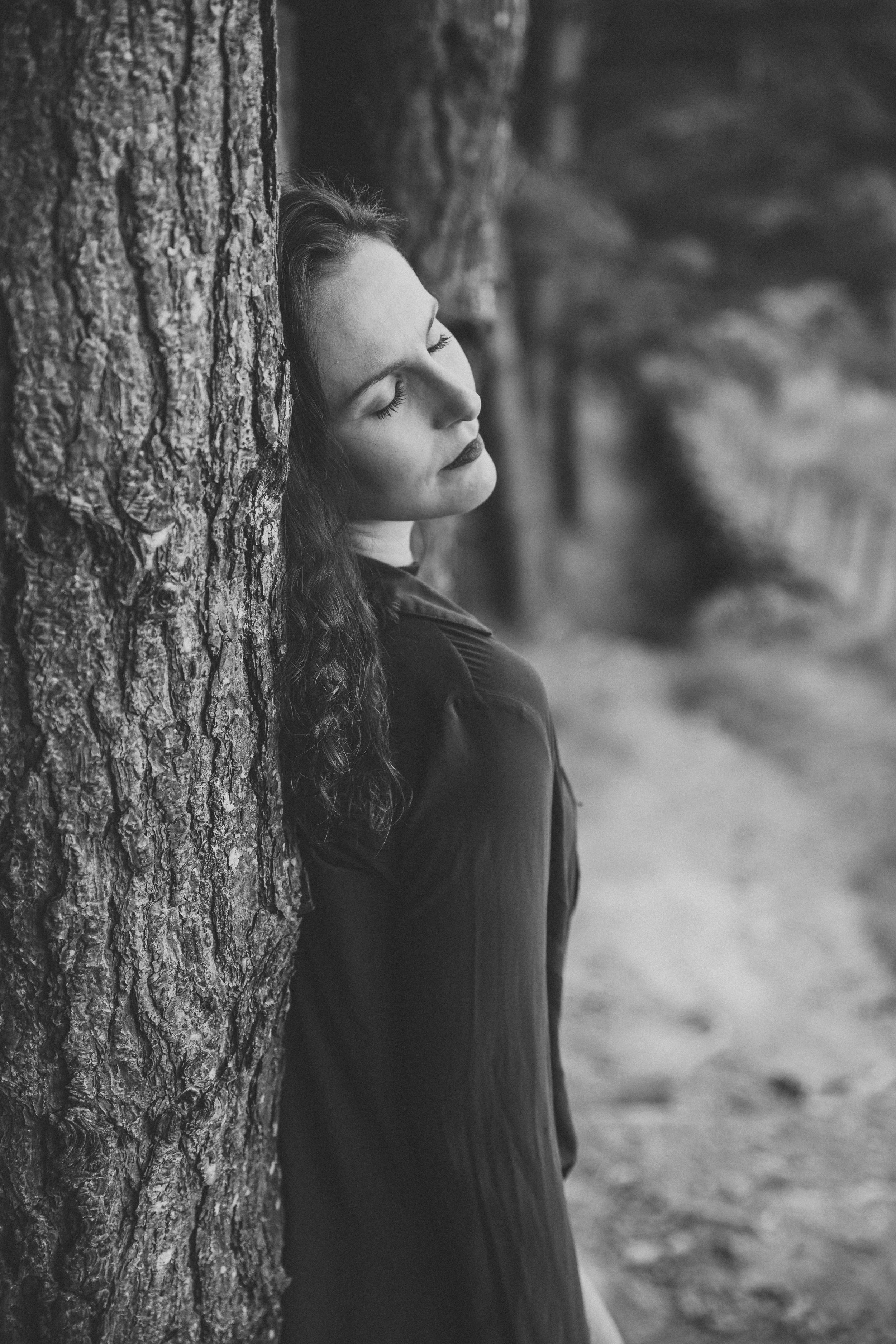 Artistic black and white portrait leaning against tree trunk in forest setting.