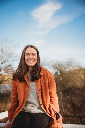 Person in orange jacket and gray sweater sitting outdoors with blue sky backdrop.