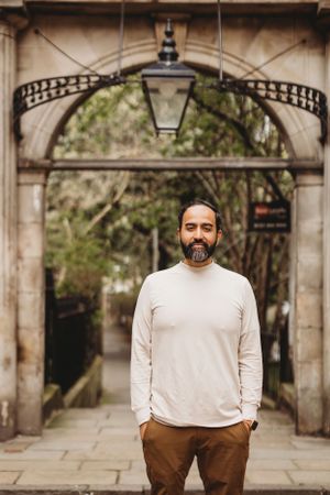 Man in cream sweater standing under ornate stone archway with lantern.