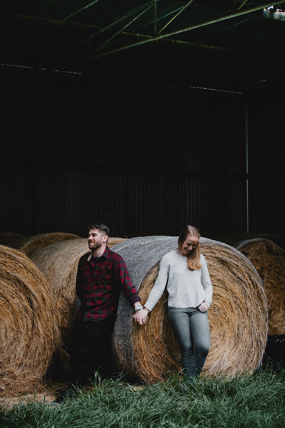 Two people lean against round hay bales in a dimly lit barn interior.