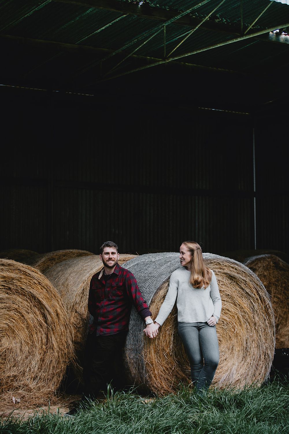 A couple poses against large hay bales in a rustic barn setting.