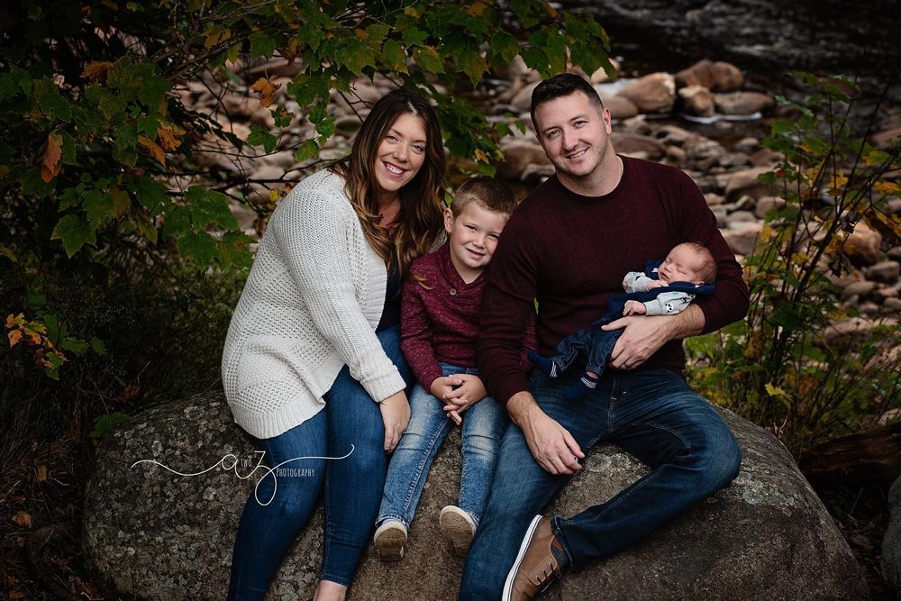 Family sitting close together on a large rock surrounded by autumn foliage.