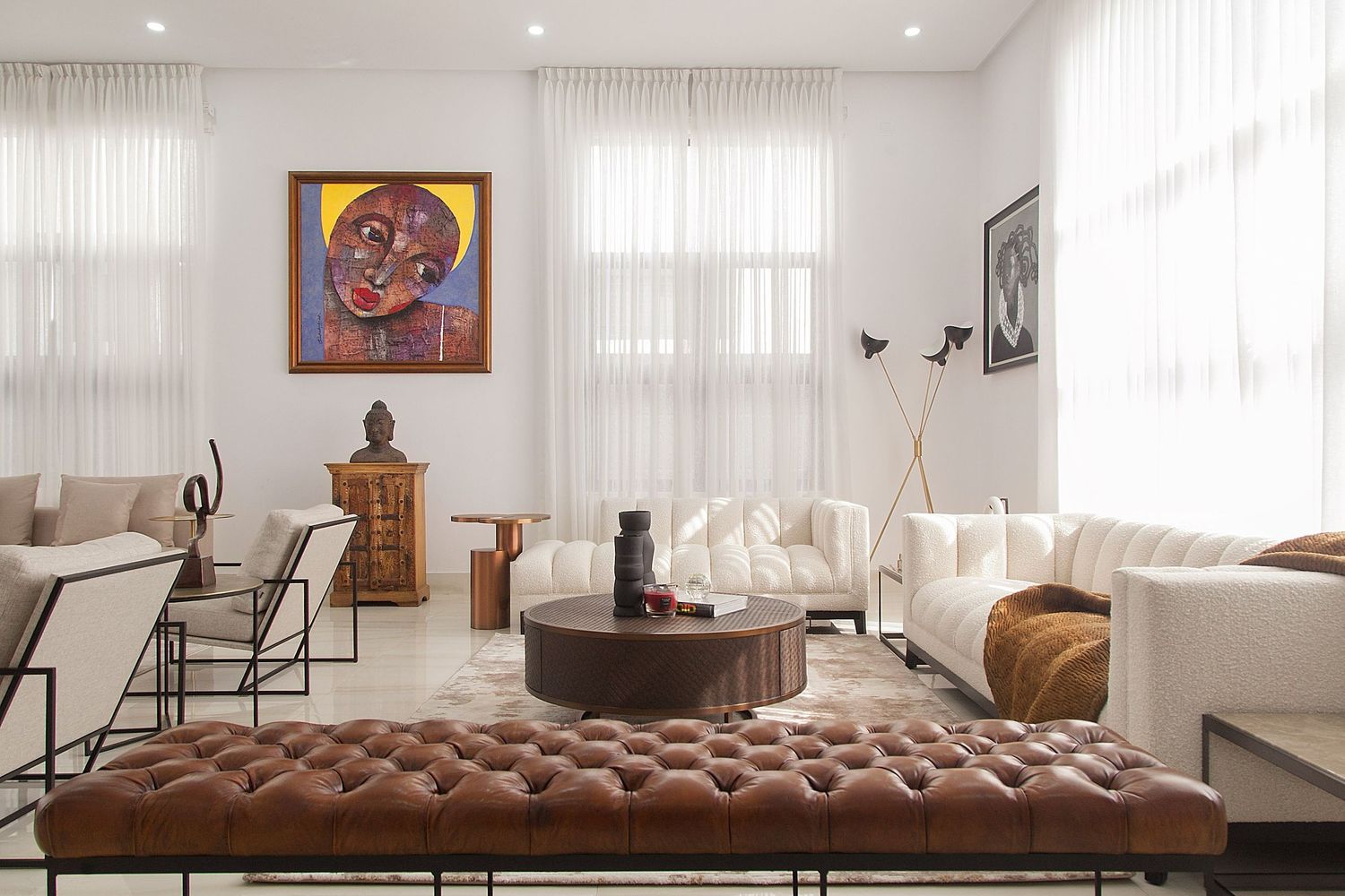 Modern living room with brown leather tufted sofa, sheer white curtains, recessed lighting and contemporary artwork.