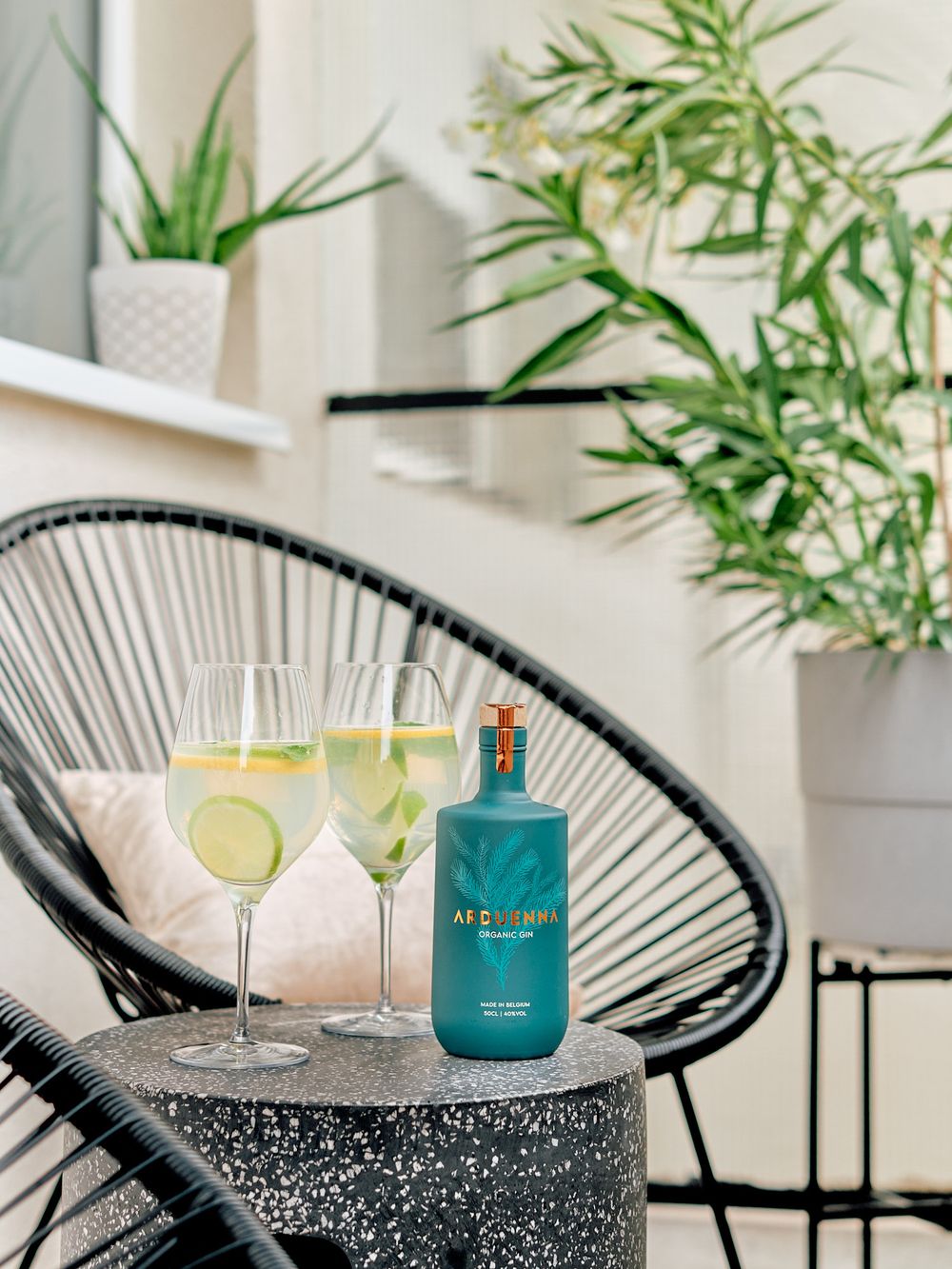 Cocktail glasses with lime garnish and blue bottle on black acapulco chair with plants.