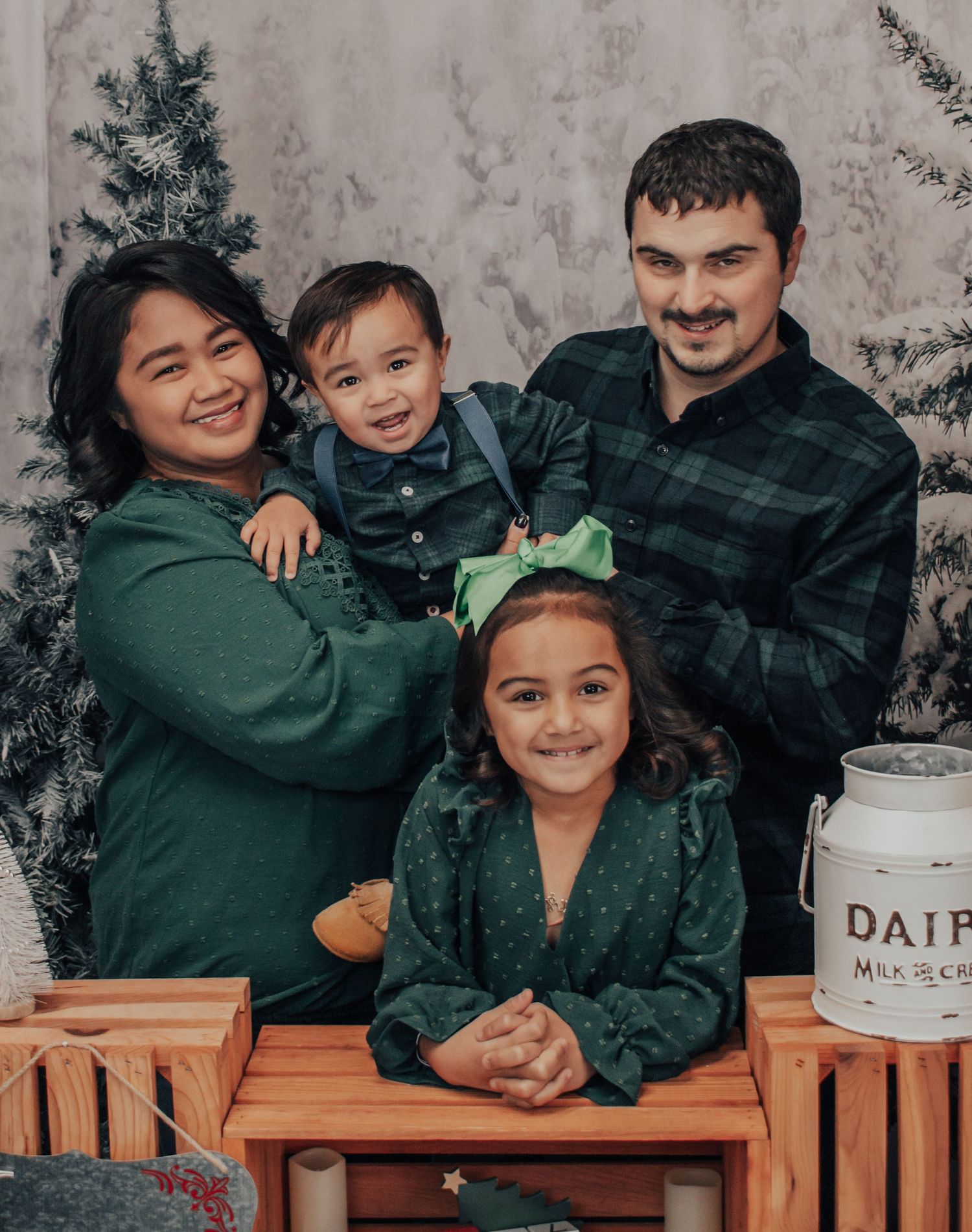 Family portrait in coordinating green outfits against textured gray backdrop with rustic wooden crates.