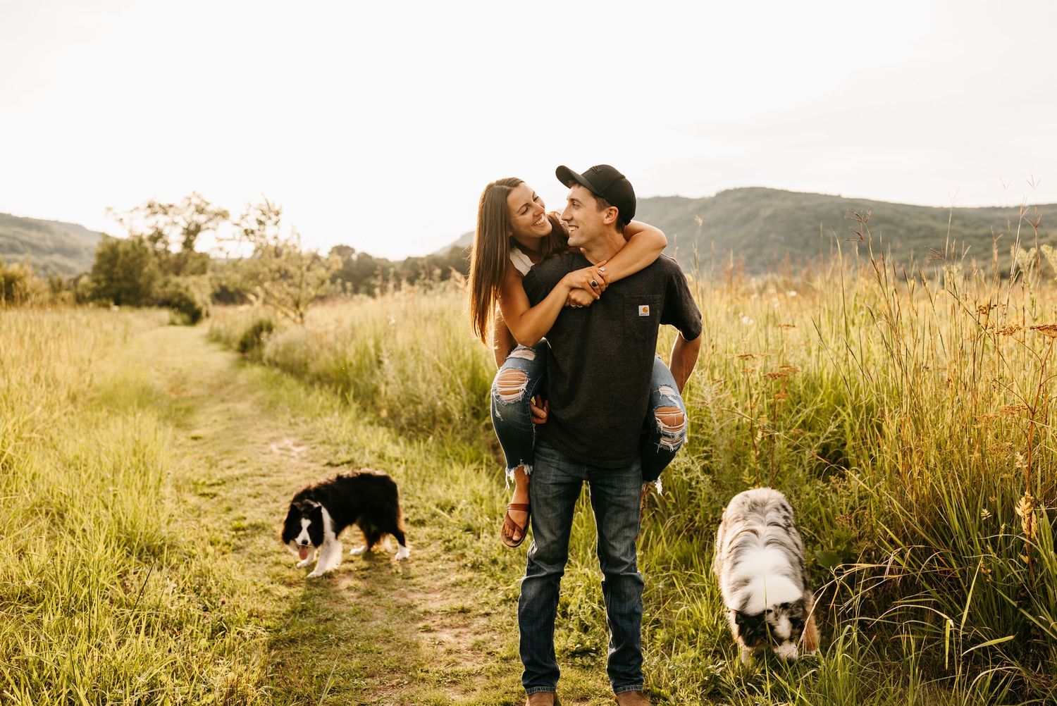 woman on back of man with arms around him looking at each other with two dogs walking next to them in grass