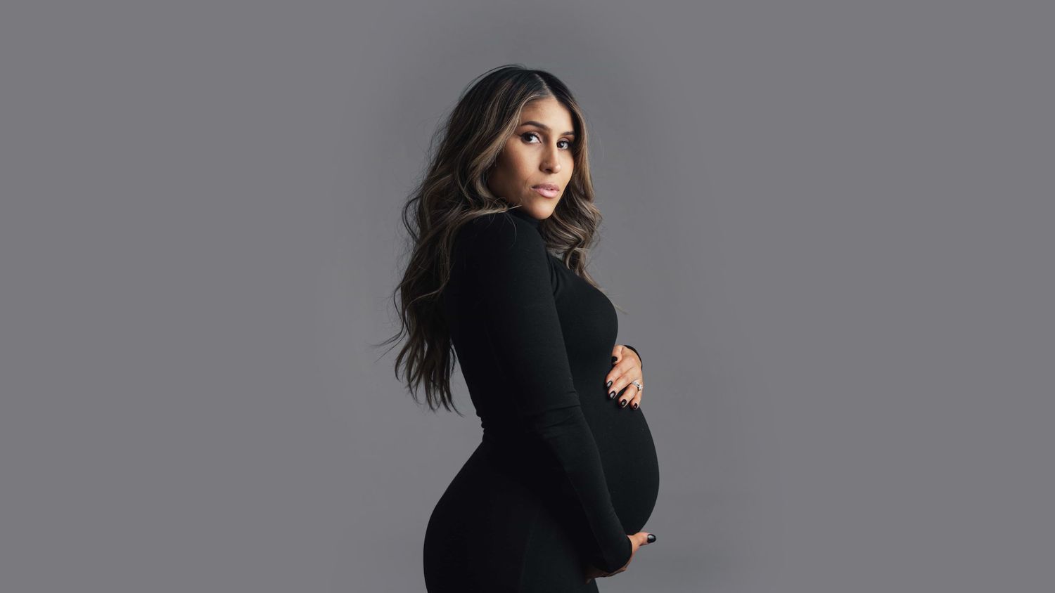 Pregnant woman in black turtleneck dress posing sideways against gray studio background.