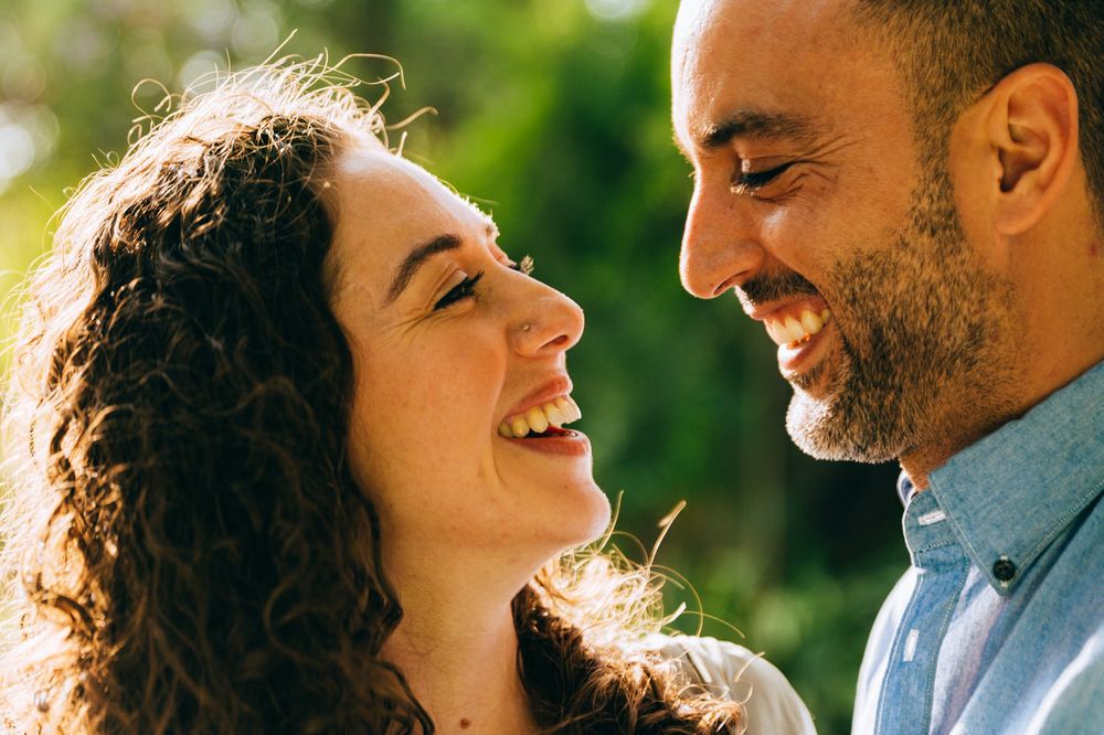 A romantic couple shares genuine laughter against sunlit greenery.