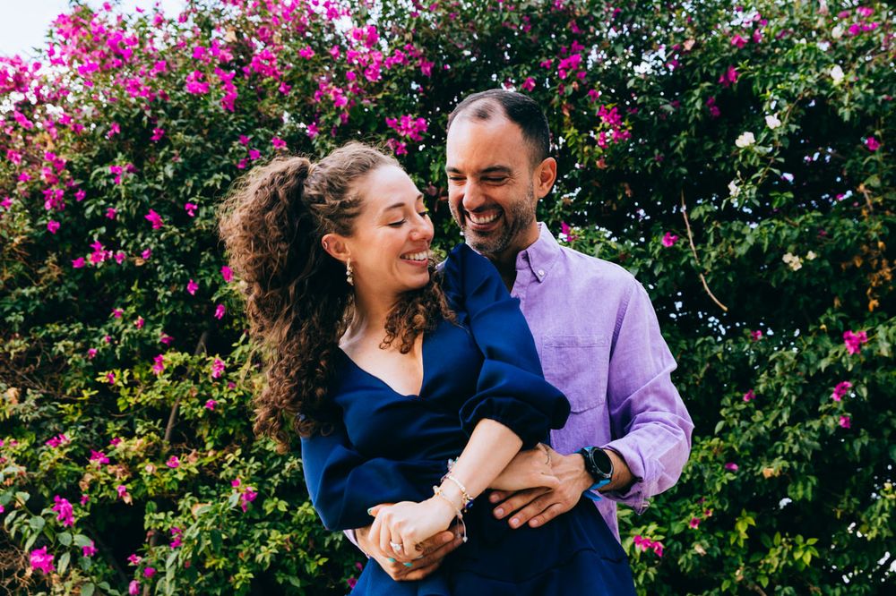 A tender embrace captured in front of pink flowering bushes on a bright day.