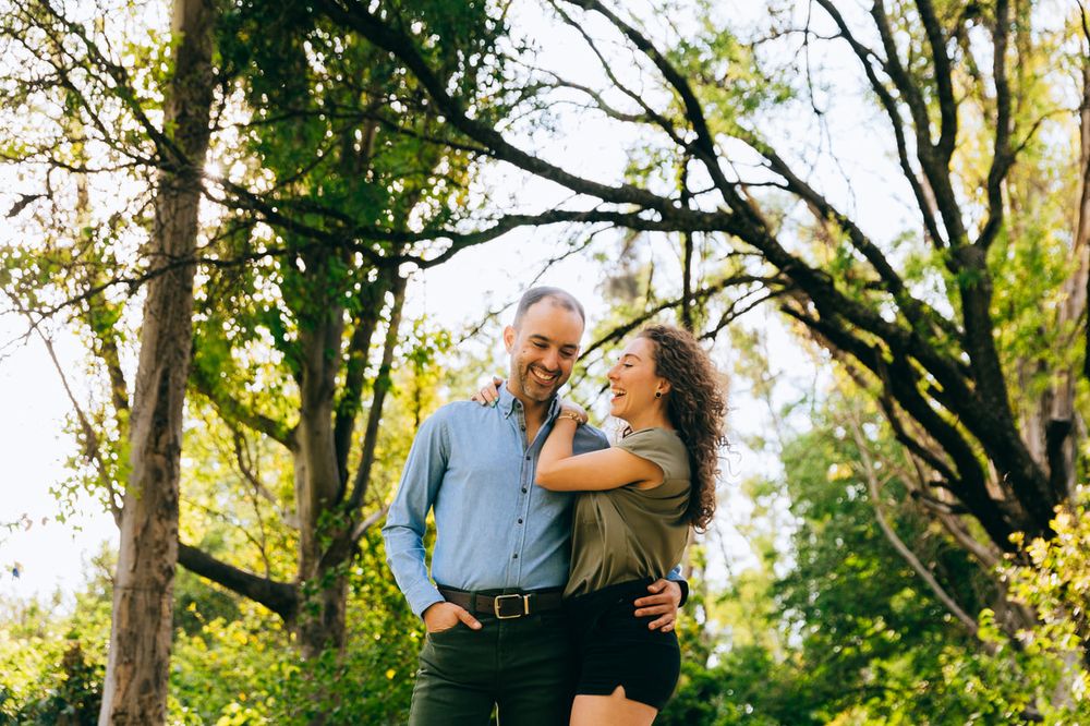 A couple laughs together while embracing under tall trees in a sunlit park.