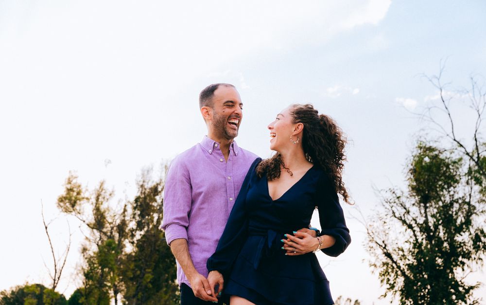 Couple laughing together outdoors in casual attire against a bright sky background.