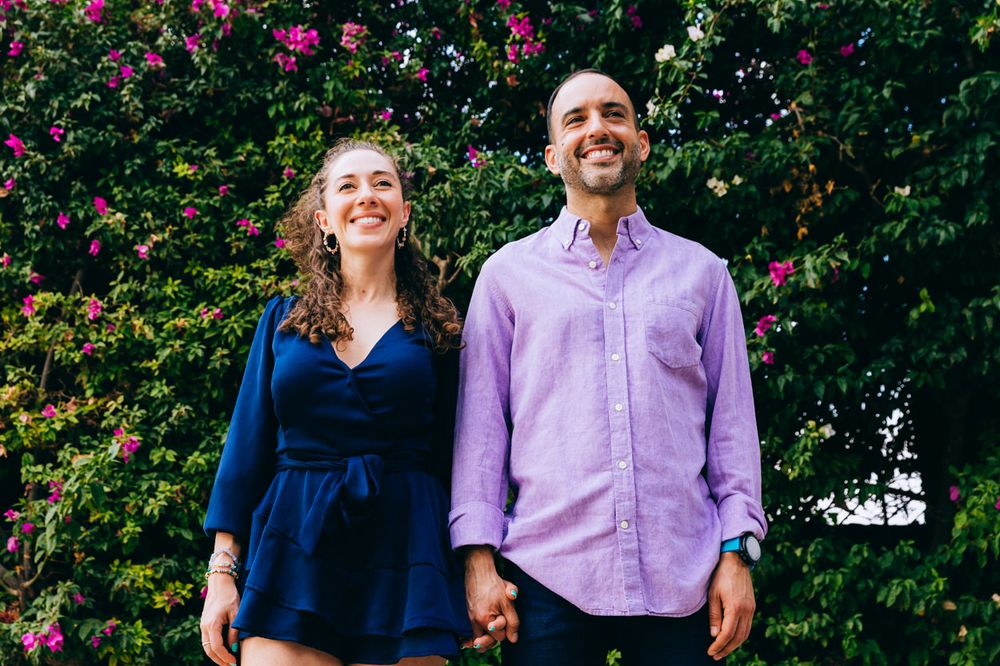 A couple stands together smiling in front of flowering pink bushes.