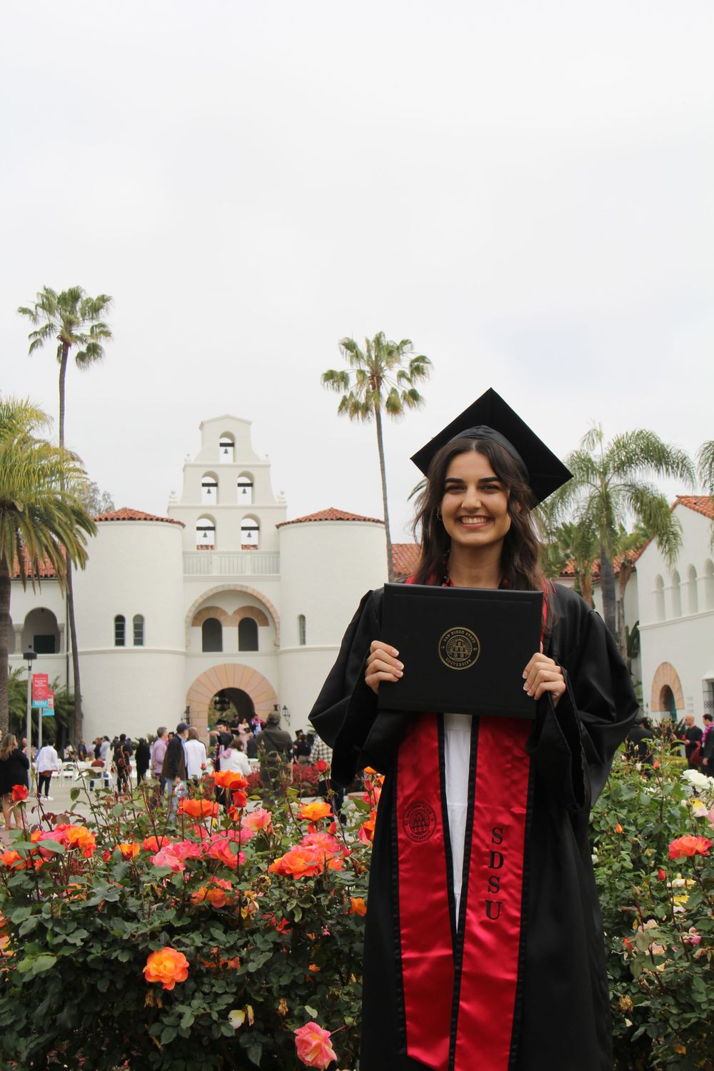 Graduate in cap and gown poses with diploma in front of Spanish mission style building with flowers.