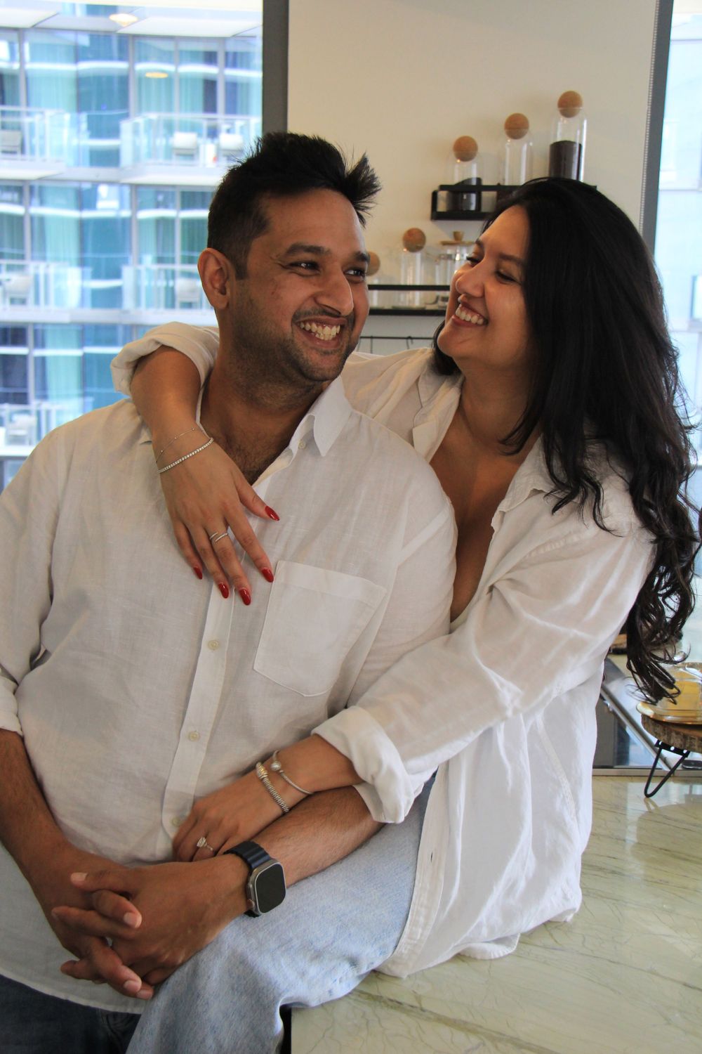 Photo by Florida photographer of a couple in white shirts embracing and laughing in a modern kitchen with Miami city views.