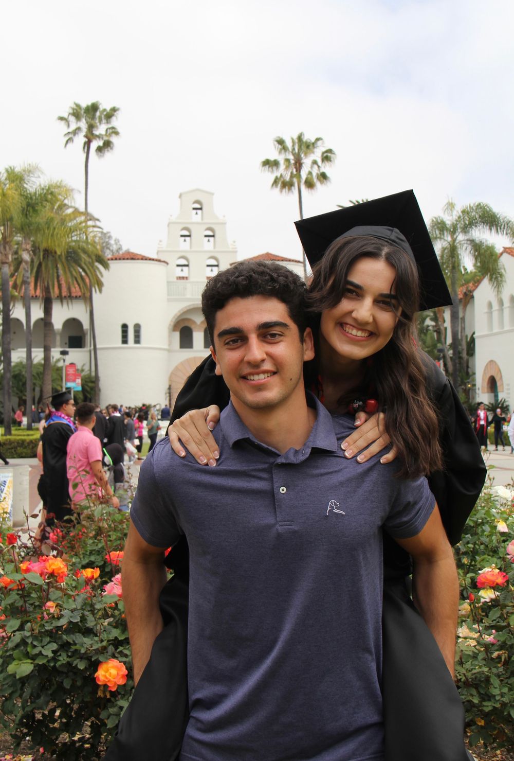 A graduation celebration photo at a Spanish-style university campus with palm trees and flowers in bloom.
