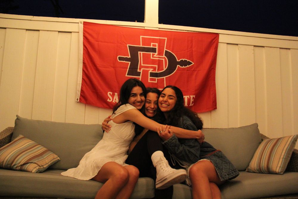 Three friends share a moment on a couch beneath a red university flag.
