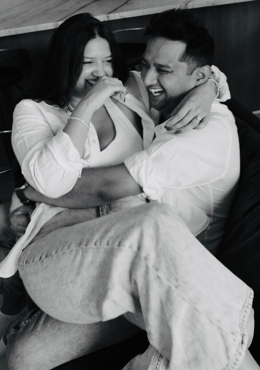 Black and white photo capturing a playful moment between a couple in casual white outfits.