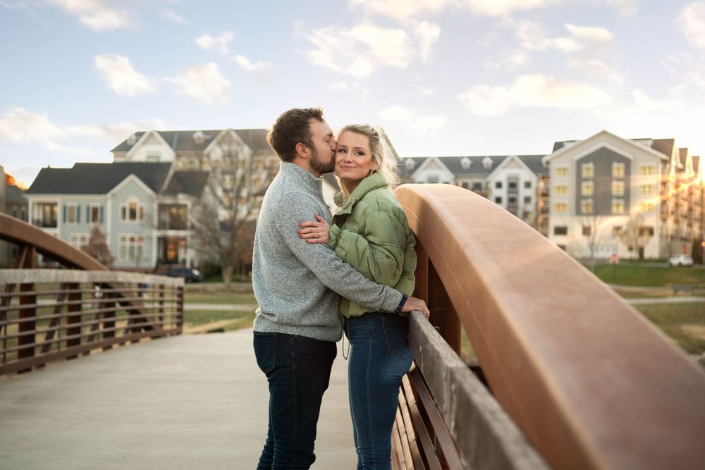 A couple sharing a romantic moment on a wooden bridge with residential buildings in the background.