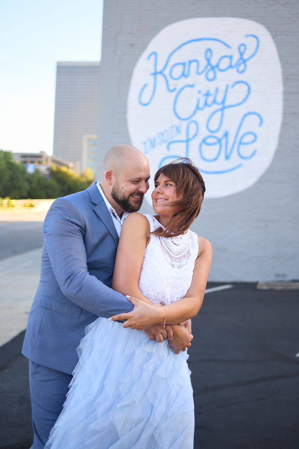 A couple embraces in front of a blue Kansas City Love mural on an urban wall.