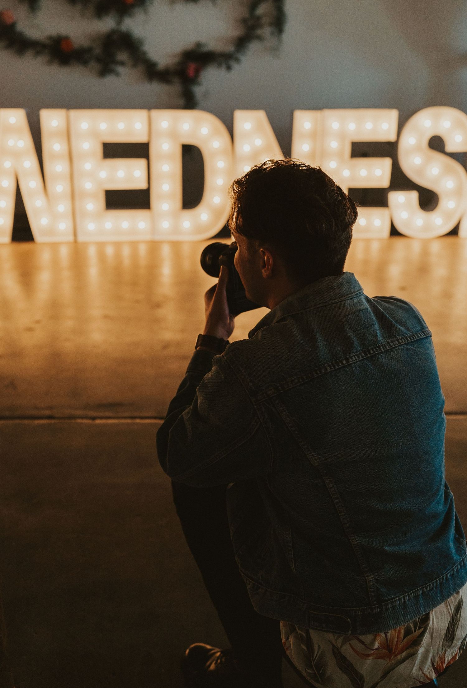 A silhouetted photographer takes a picture in front of illuminated 'WEDNES' marquee letters creating a moody atmosphere.
