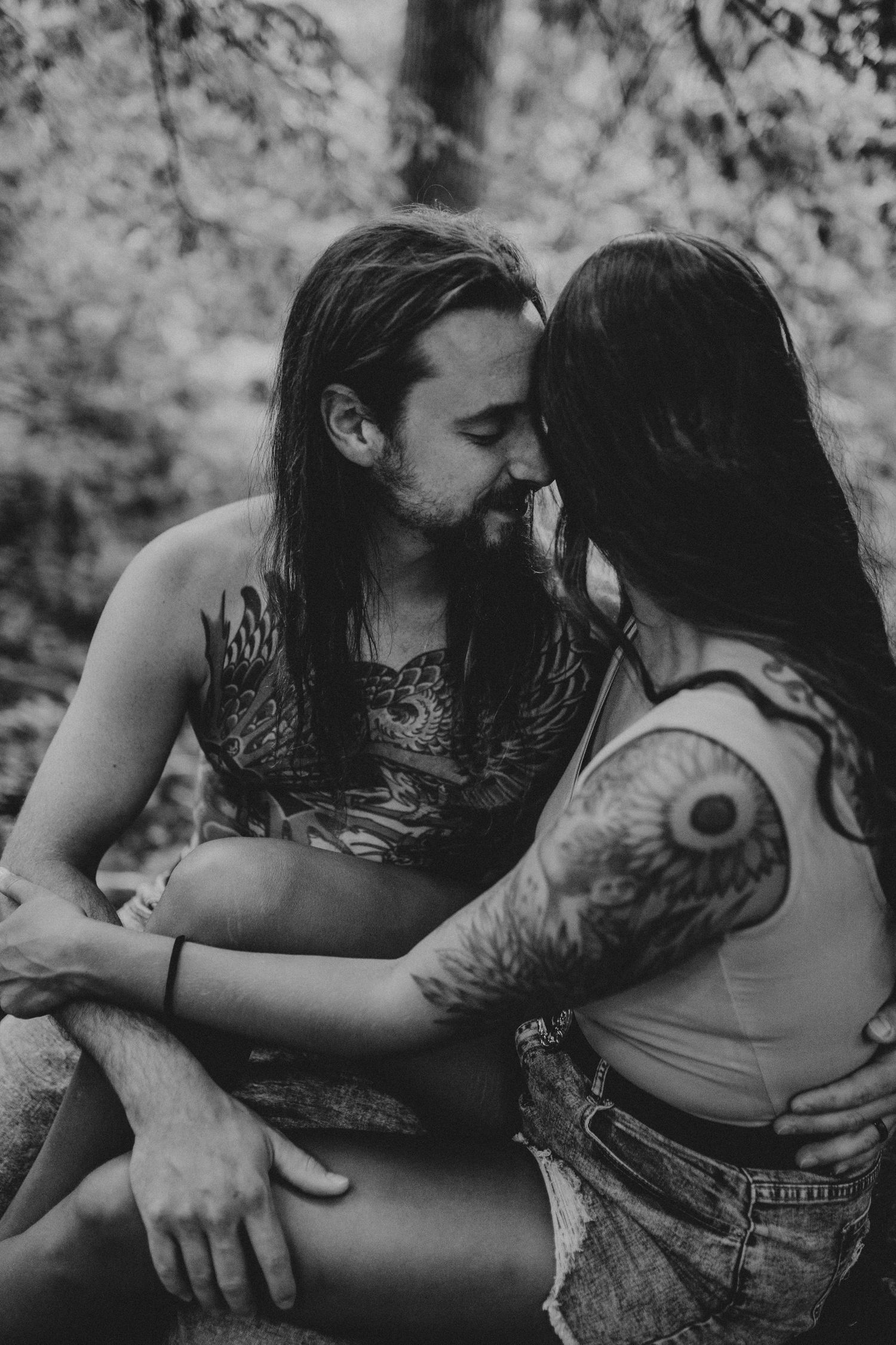 black & white photo of a tattoo-covered couple snuggled up together sitting on a boulder in the forest