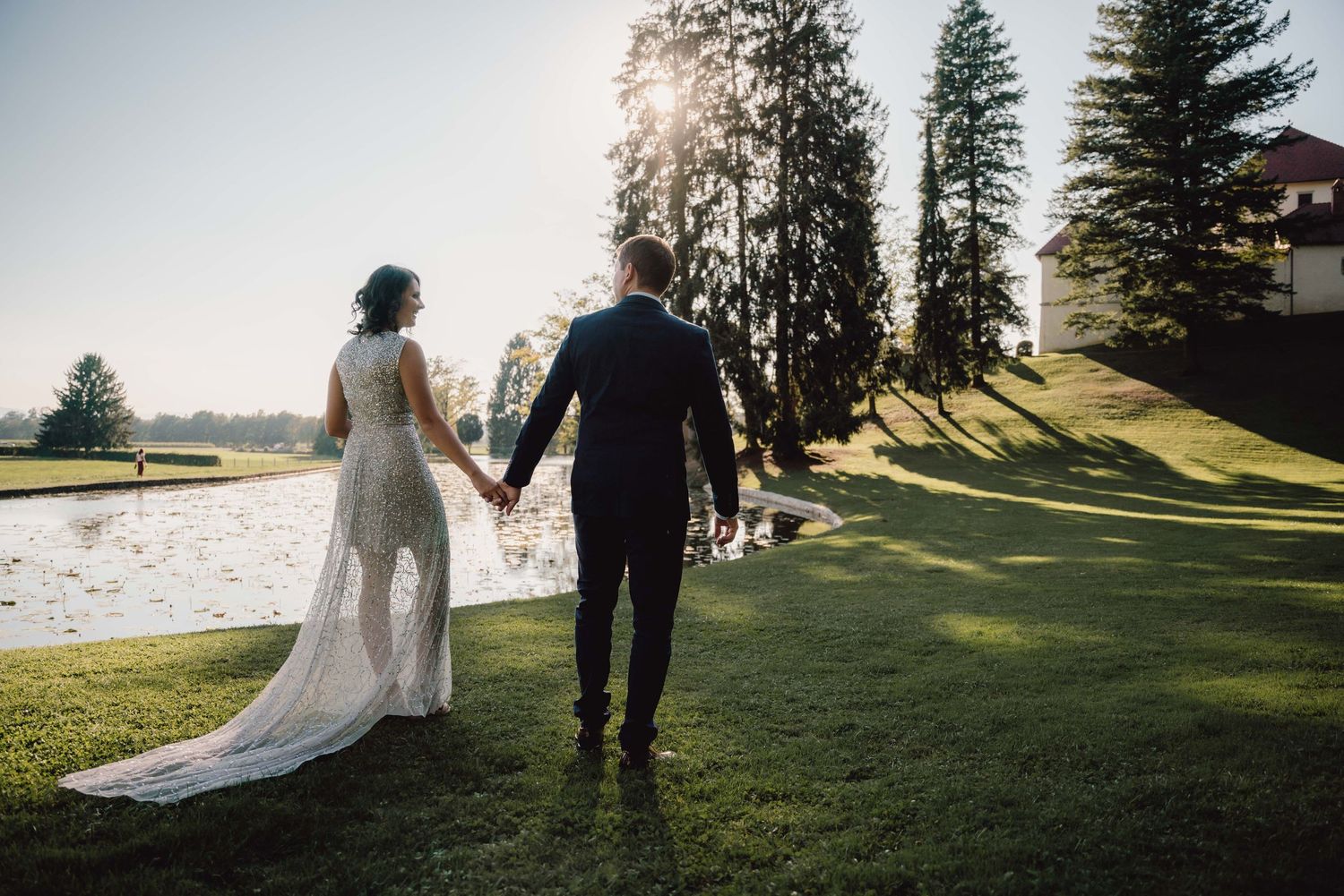 A couple holds hands while walking on a grassy hill near evergreen trees during golden hour.