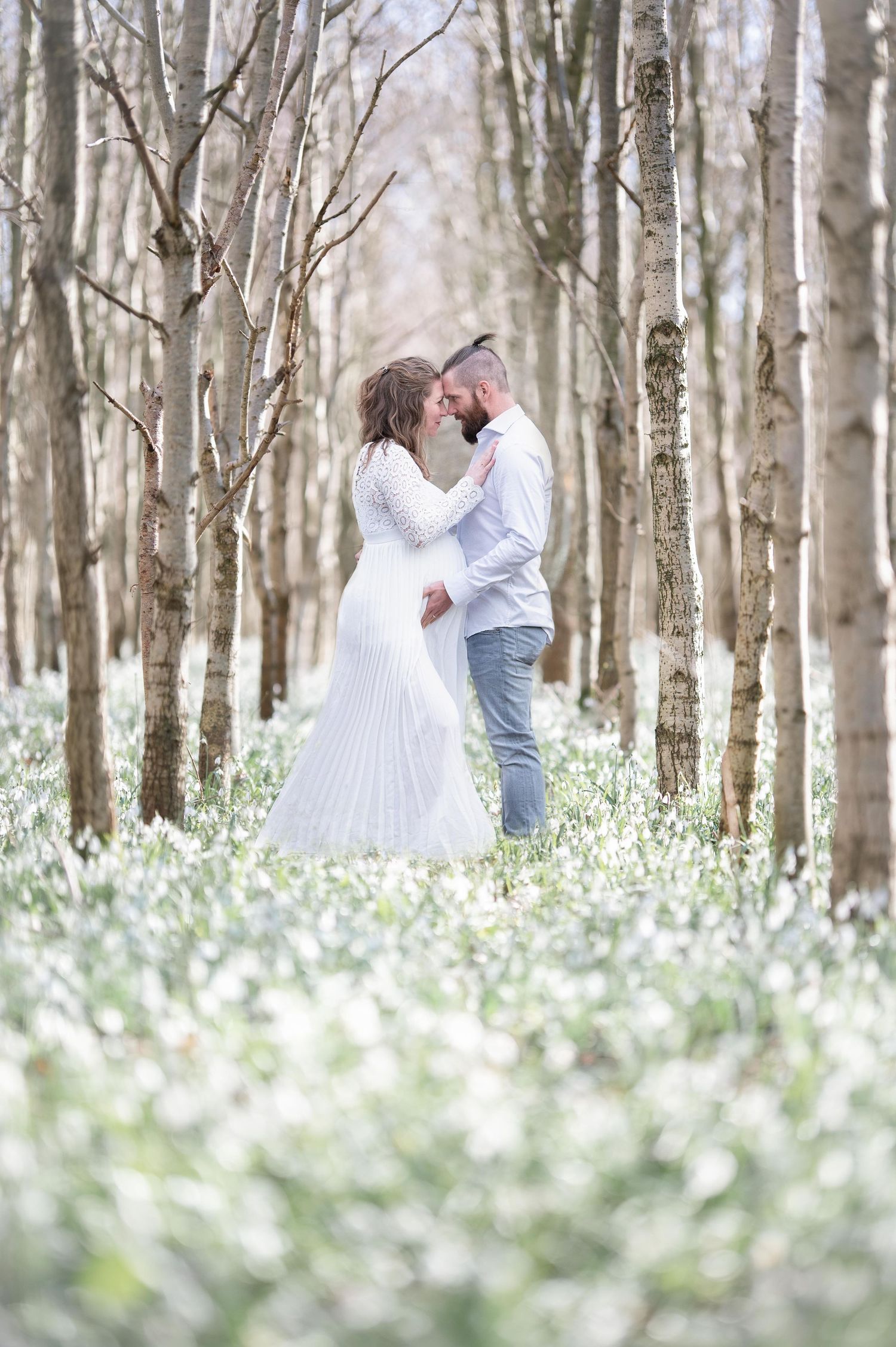 zwangere vrouw en partner staan tussen bloeiende sneeuwklokjes en kijken verliefd naar elkaar met zijn hand op haar buik
