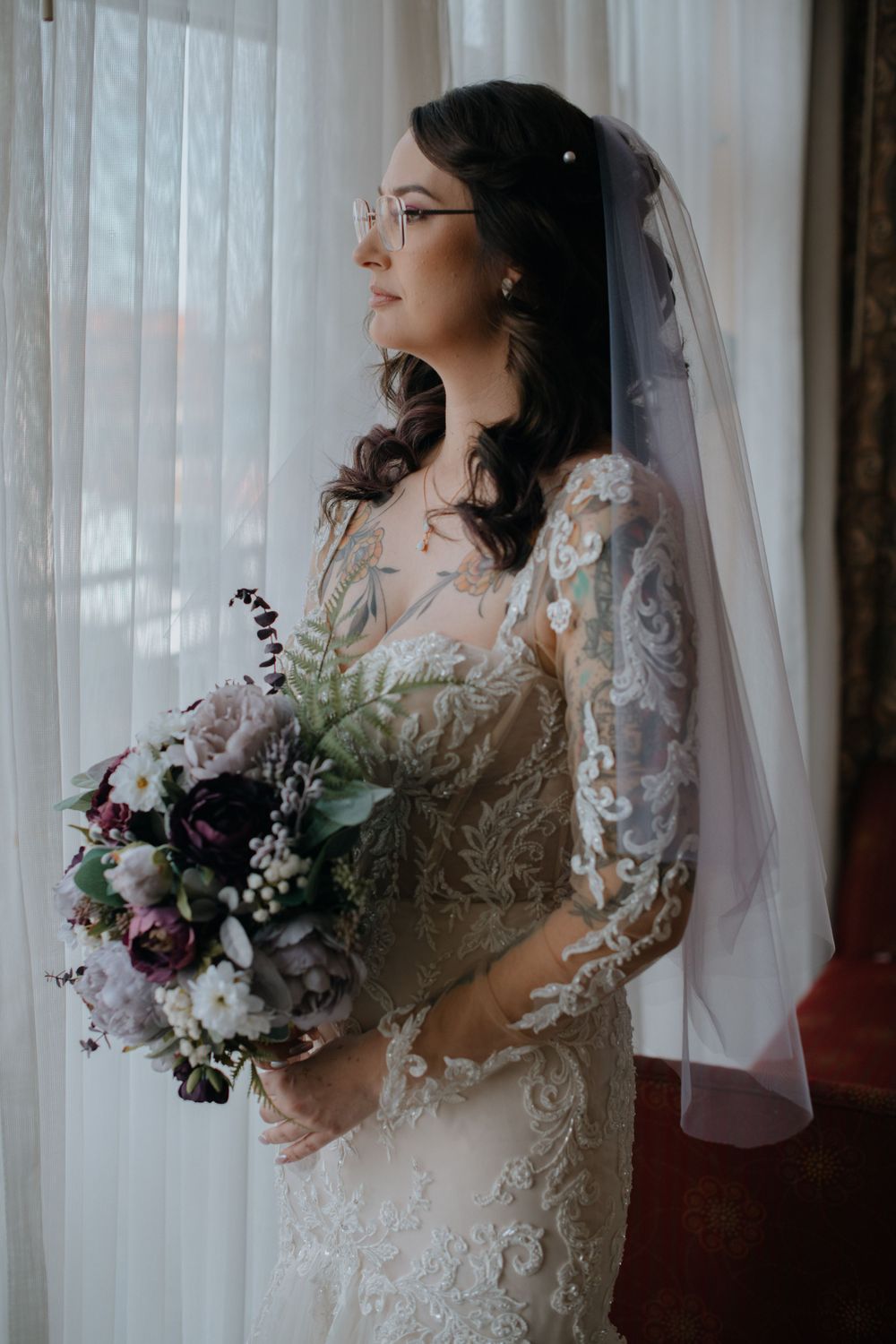 Bride in lace dress with glasses holds purple and white bouquet by window with sheer curtains.
