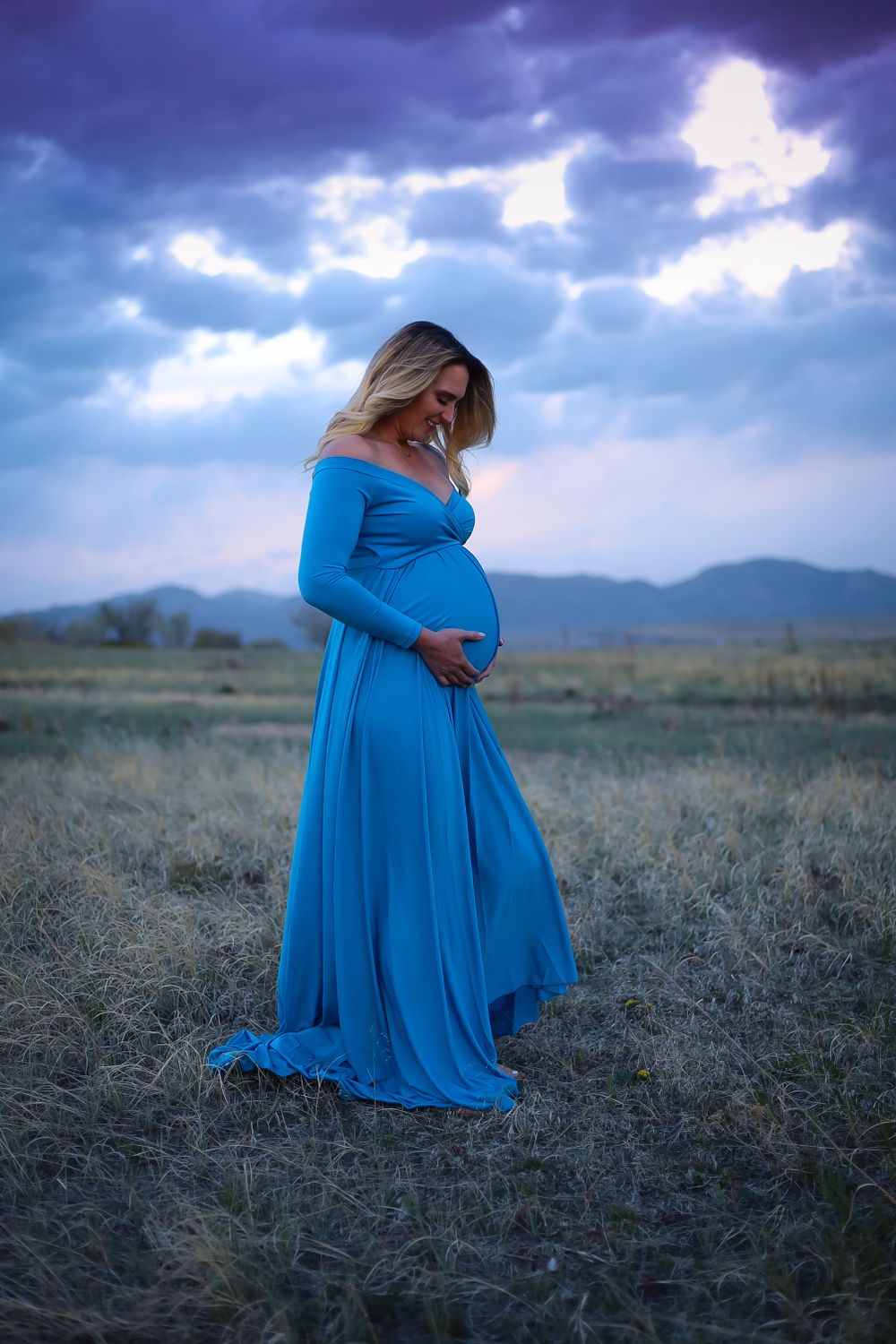 A dramatic maternity portrait in a flowing blue dress against a stormy mountain landscape.