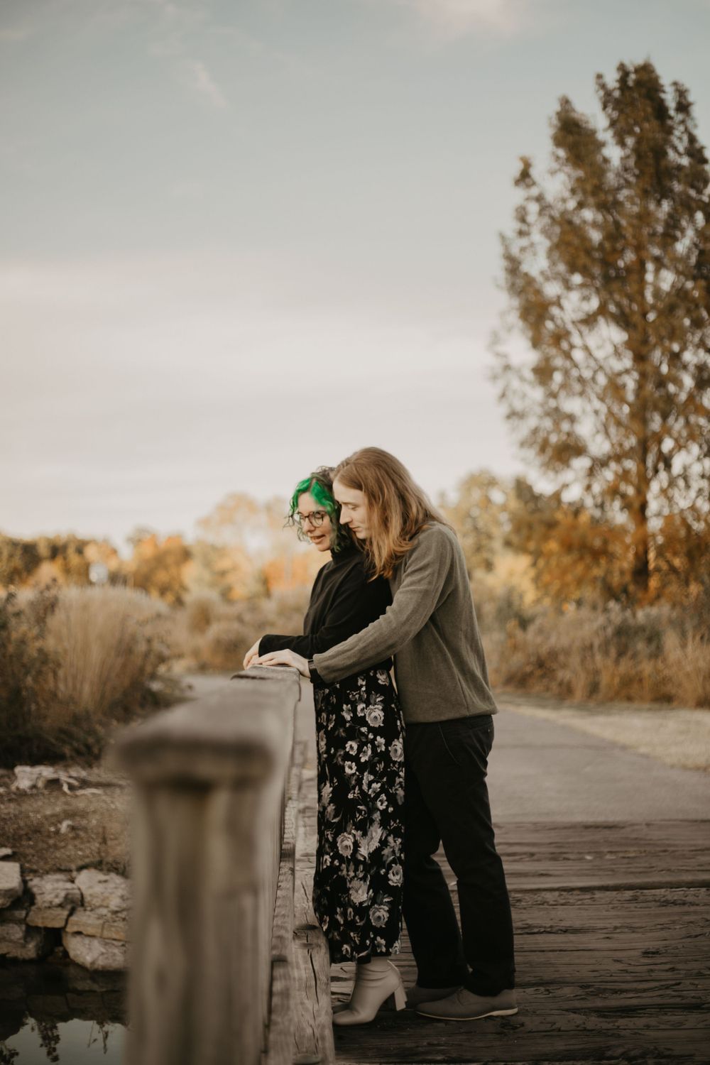 Couple leaning on railing during golden hour with autumn trees in background.