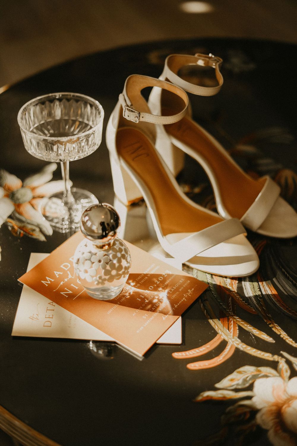Wedding accessories including white heels, crystal glass, and invitation cards arranged on dark surface.