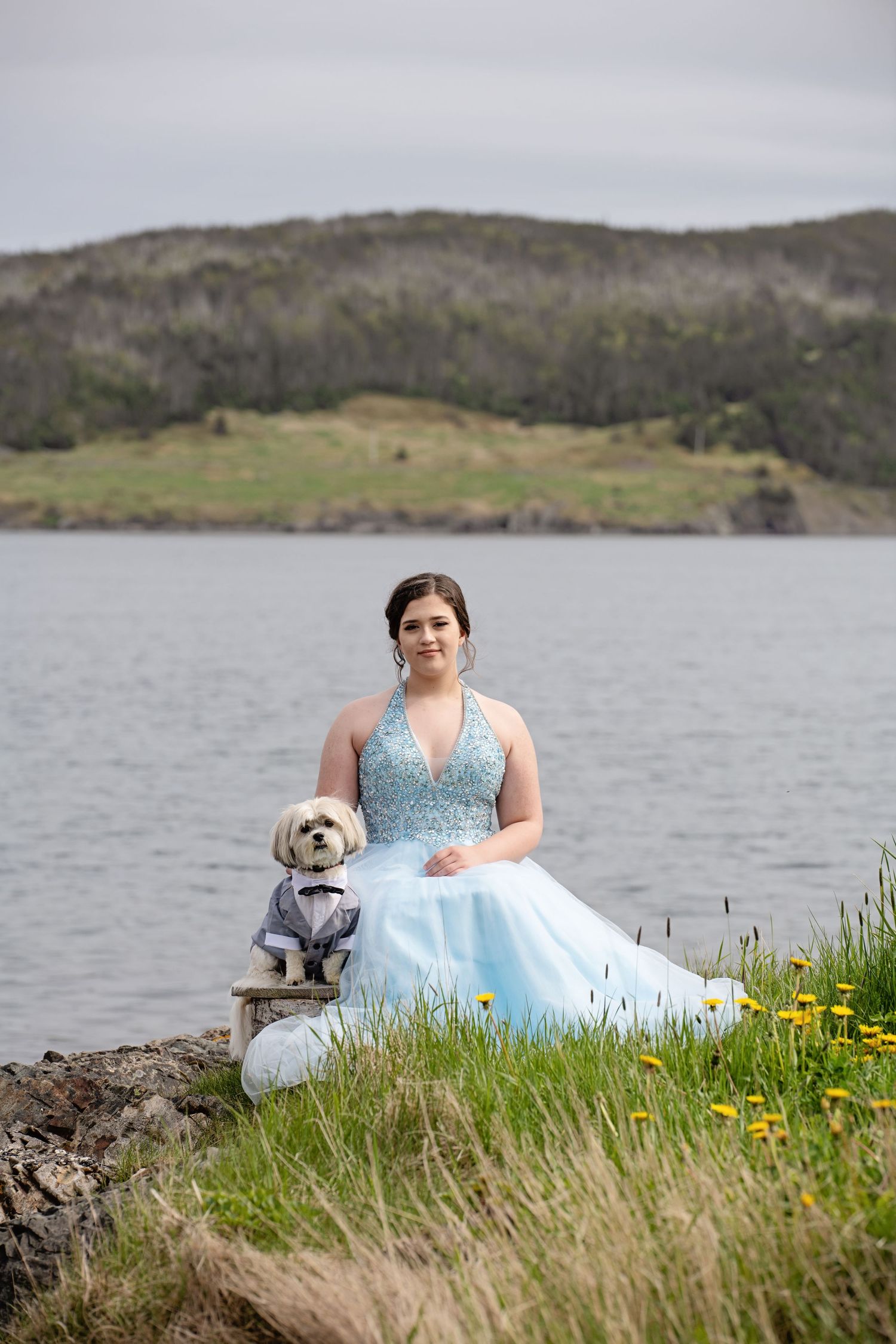 Person in blue gown sitting with small dog by lake with hills in background.