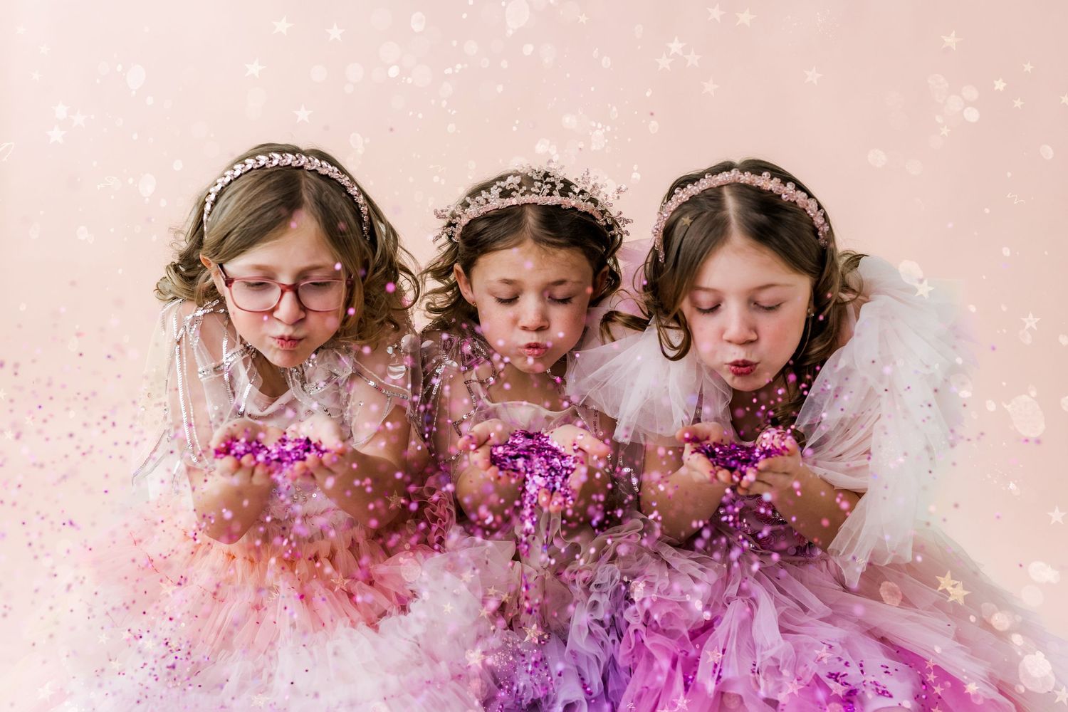 three little girls dressed in dresses and tiaras blowing glitter
