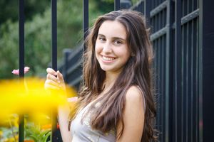 Flowers provide some noise in the foreground of this image. A beautiful girl hangs on a black iron fence and smiles