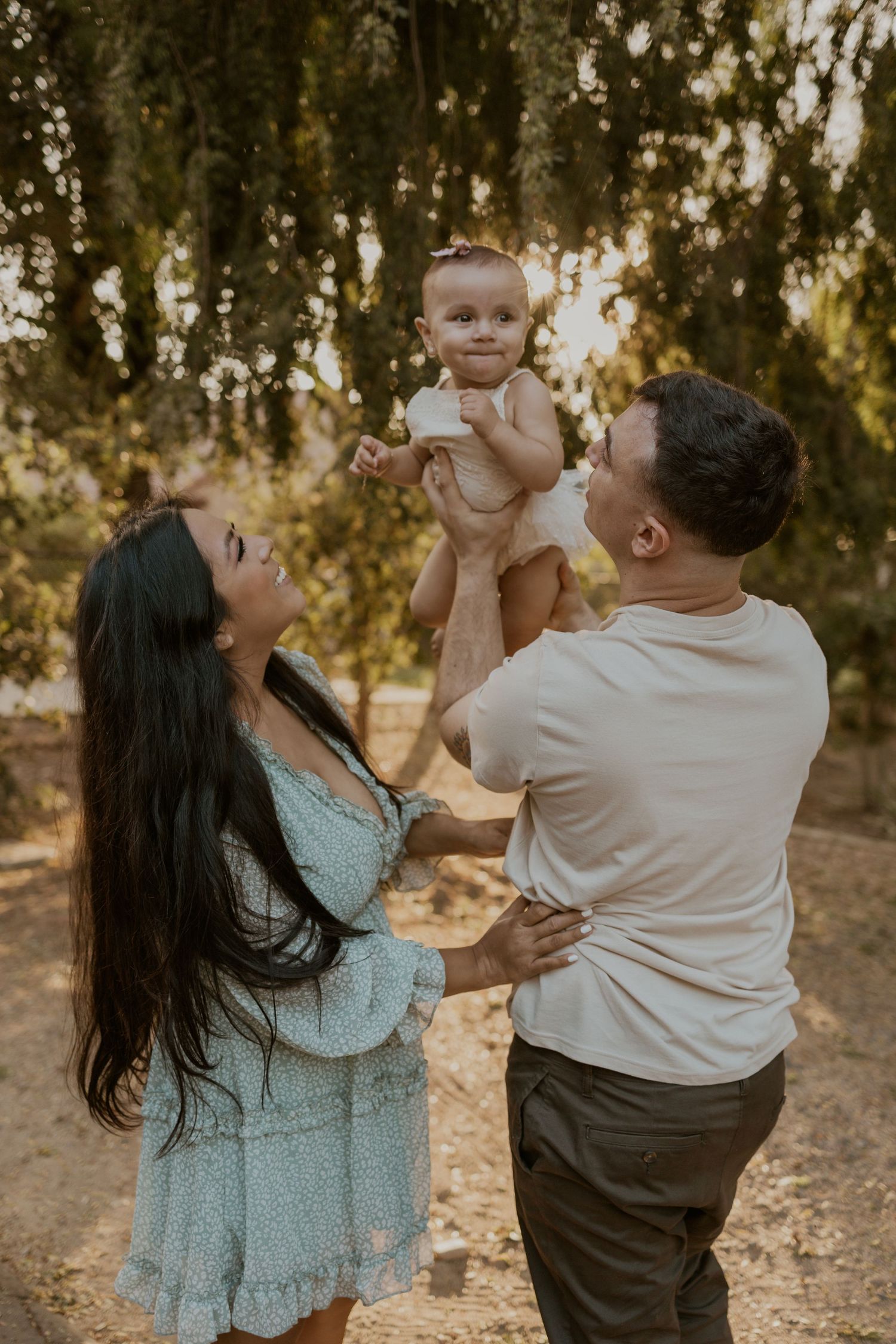 A man holds a baby up while a woman smiles at them, with trees and sunlight in the background.