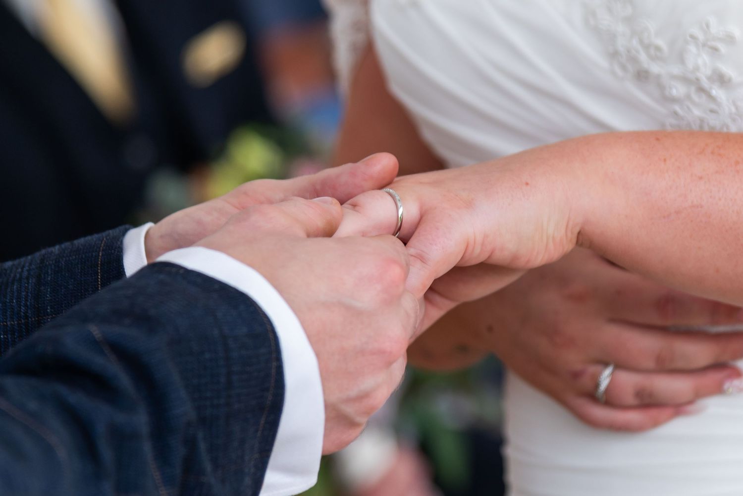 Close-up of wedding rings being exchanged during ceremony with hands visible against white backdrop.