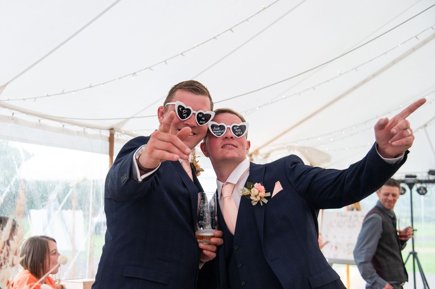Wedding guests wearing sunglasses celebrate under a white marquee tent.