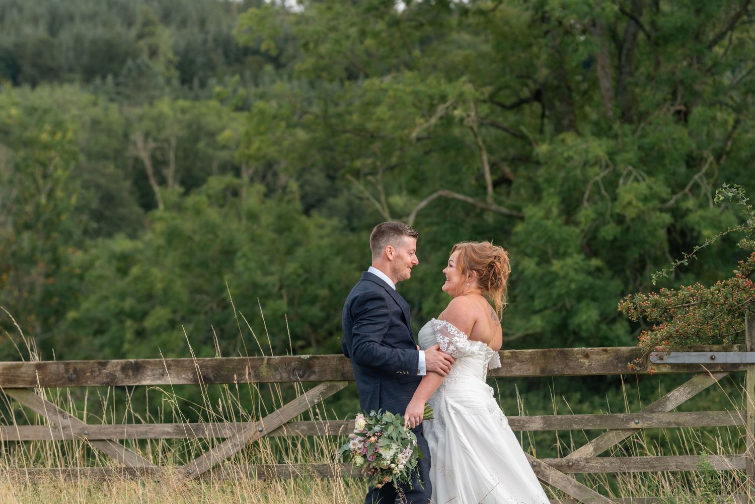 Wedding couple poses in romantic natural outdoor setting with rustic wooden fence and lush greenery background.